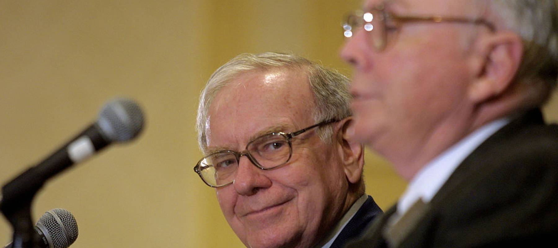 Warren Buffett seen in background but with camera focus smirking, while Charlie Munger is seen from the side, out of focus, speaking.