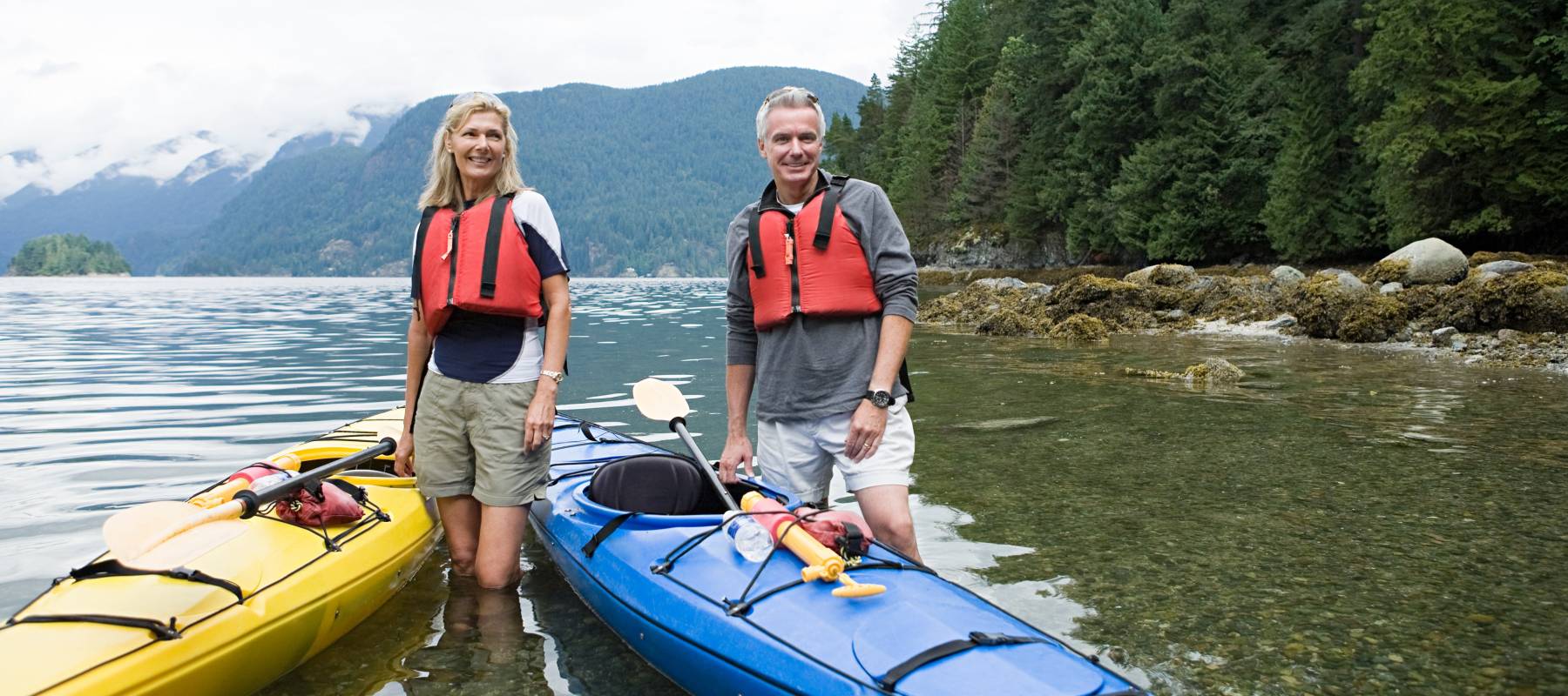 Older couple about to set out on a kayak ride, wearing life jackets and smiling for the camera standing in water.