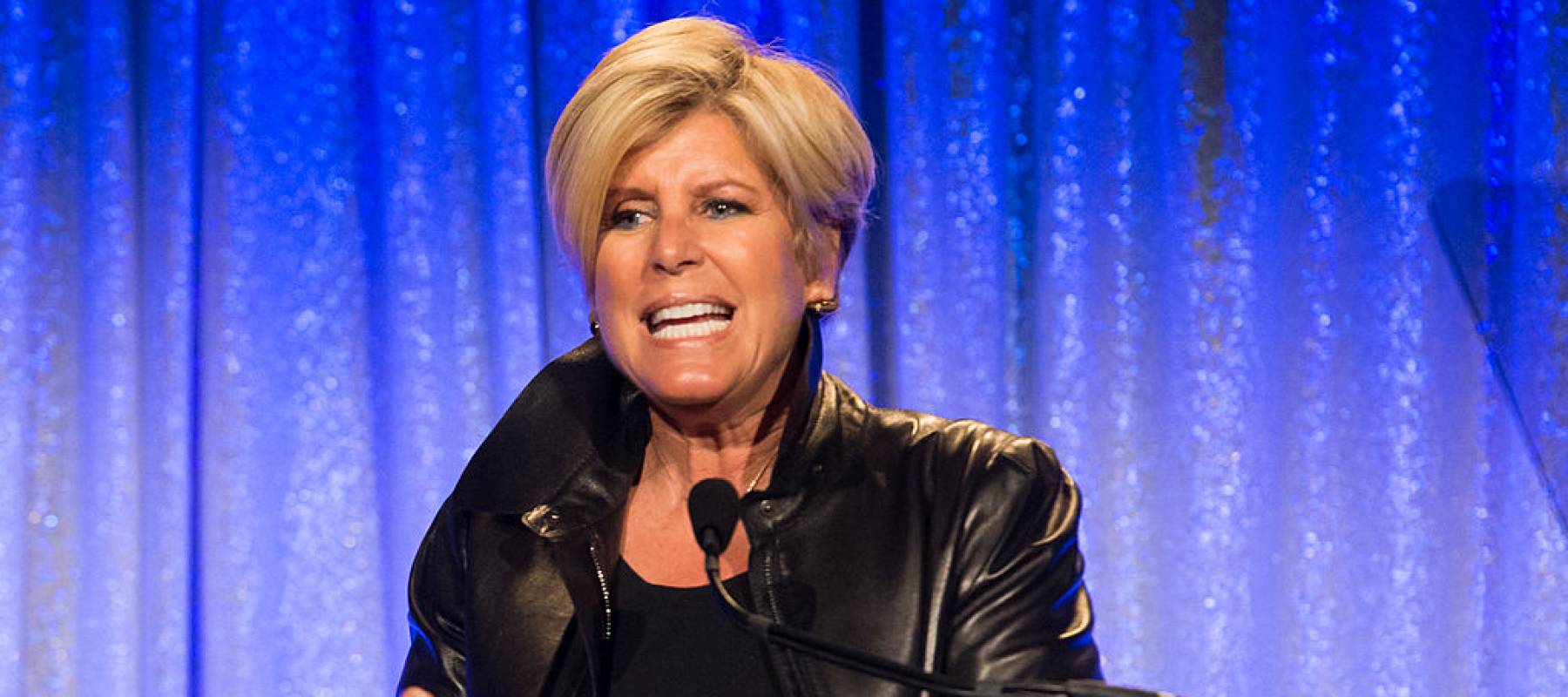 Suze Orman speaks vehemently on stage in front of a blue backdrop.