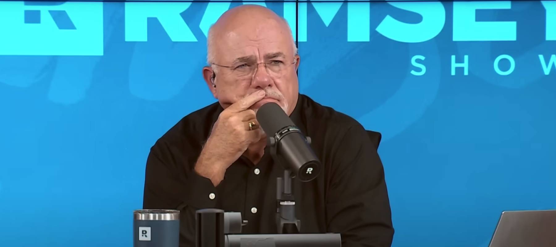 Dave Ramsey speaks into his microphone on set of the Ramsey Show, listening with his pointer finger on his lips in a thoughtful look.