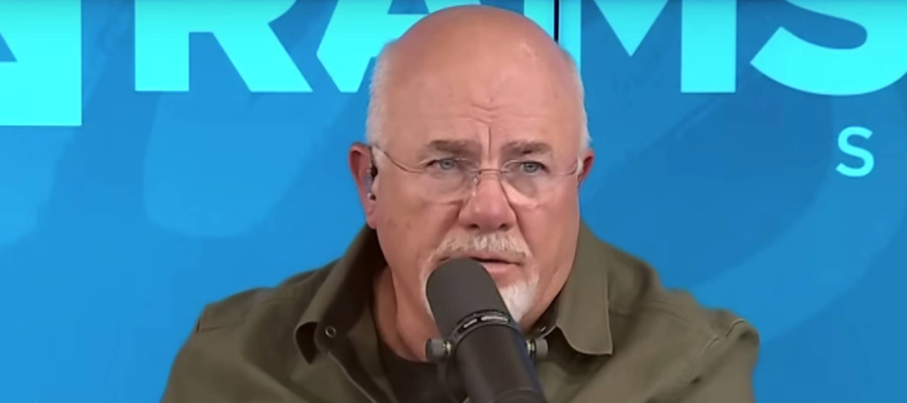 Dave Ramsey makes a dramatic face while speaking into microphone on set of his podcast.