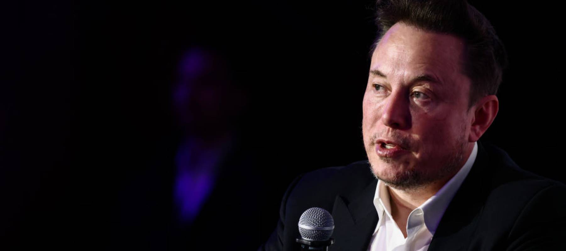 Elon Musk seen on stage, sitting in darkness, looking serious.