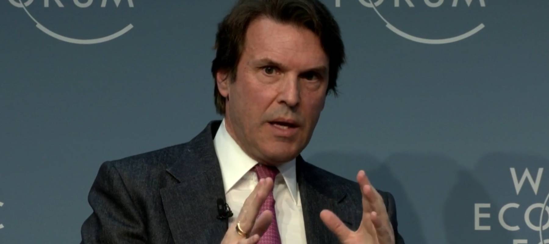 Man in suit, speaking with his hands held up while sitting in front of a World Economic Forum backdrop.