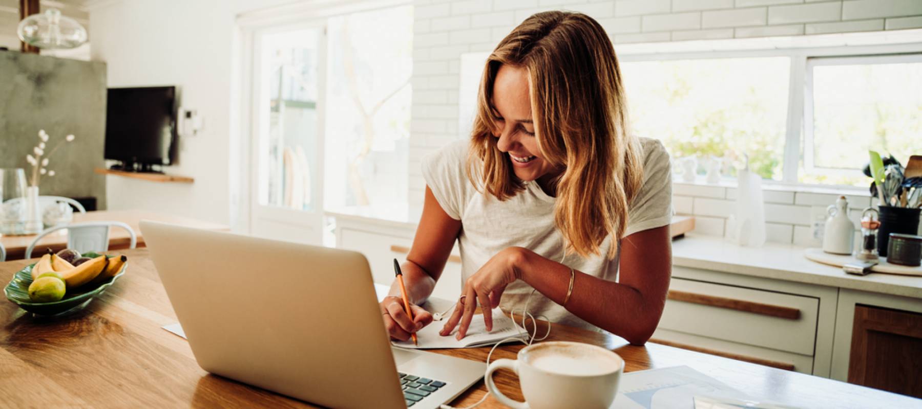 Caucasian female student working from home writing in note pad with laptop sitting next to hot coffee