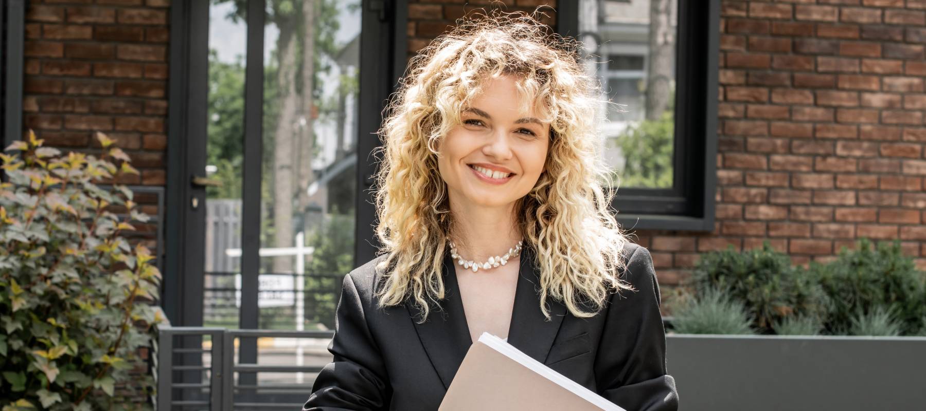 Stylish blonde real estate agent looking at documents in folder near contemporary urban building.
