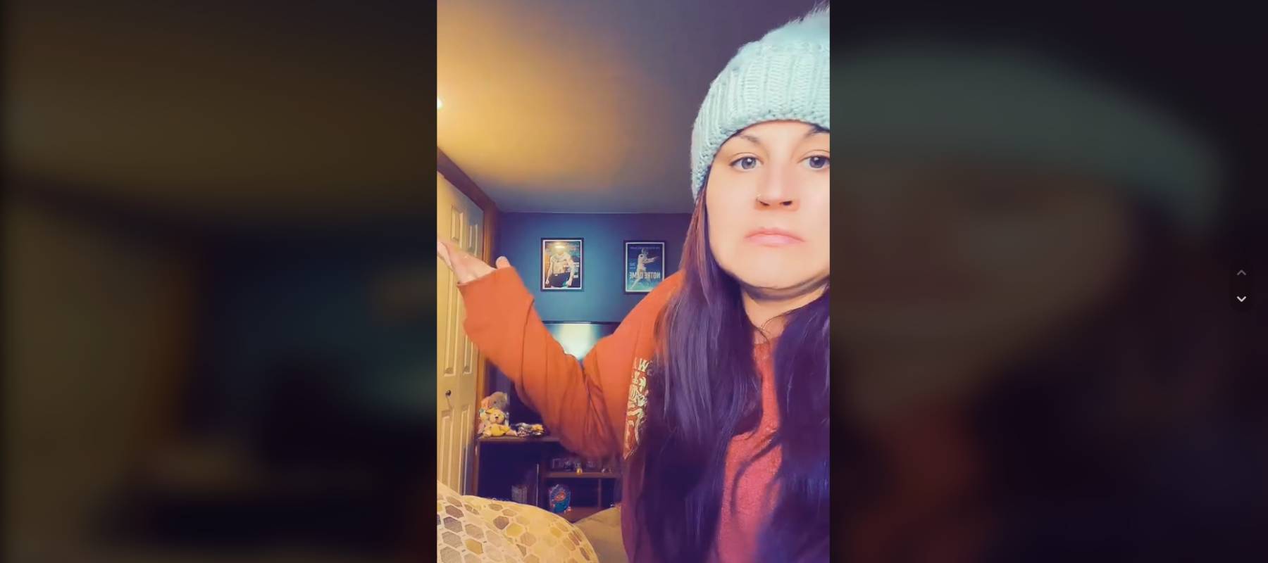 Woman wearing a knit hat in a bedroom, posing with her arm up in a shrugging gesture.