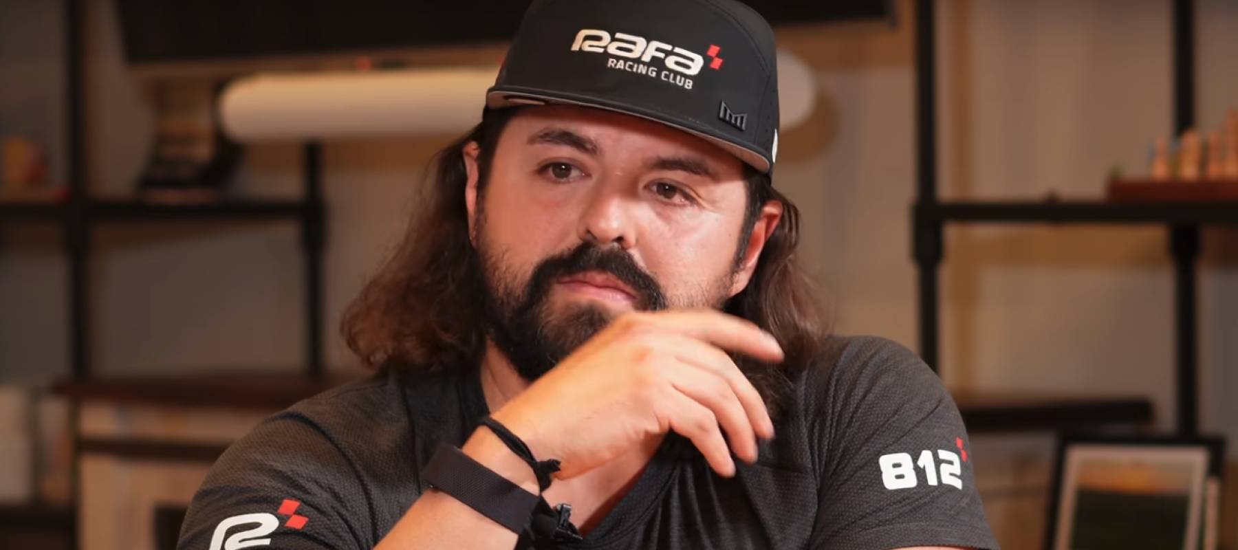 Man with beard wearing black baseball hat, talking and holding hand up to face.