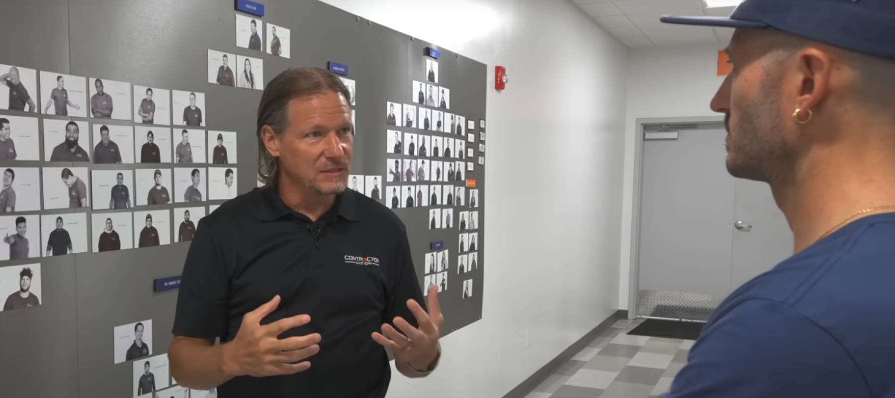 Two men speaking, facing each other in front of a wall with photos of headshots lined up.