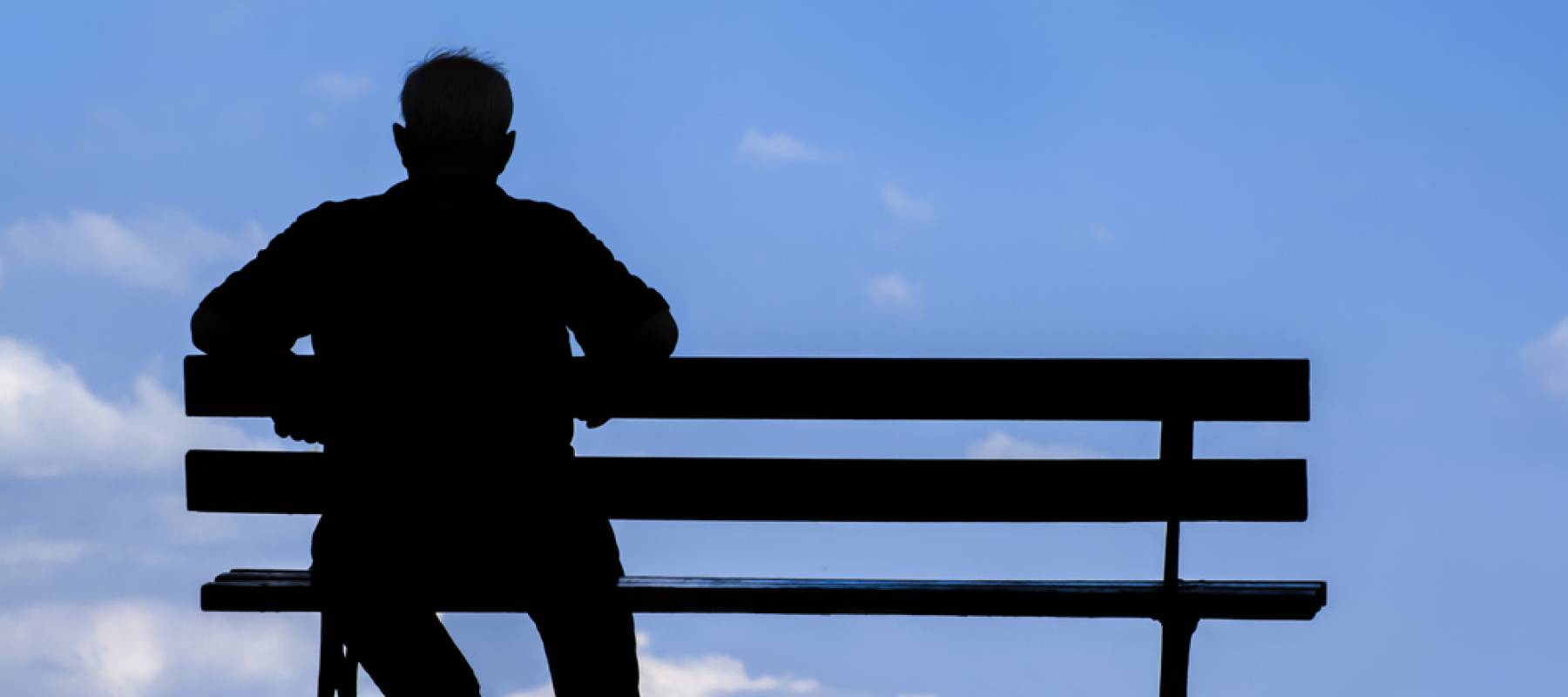 silhouette of old man, with back to camera, sitting alone on park bench under tree