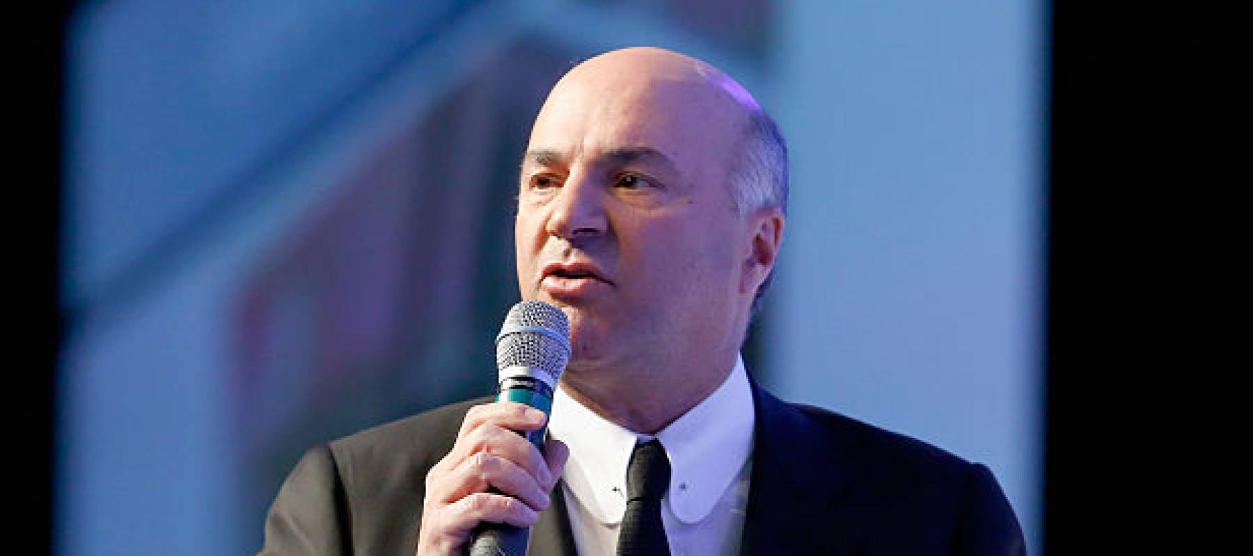 Kevin O'Leary seen on stage talking into a microphone, looking serious.