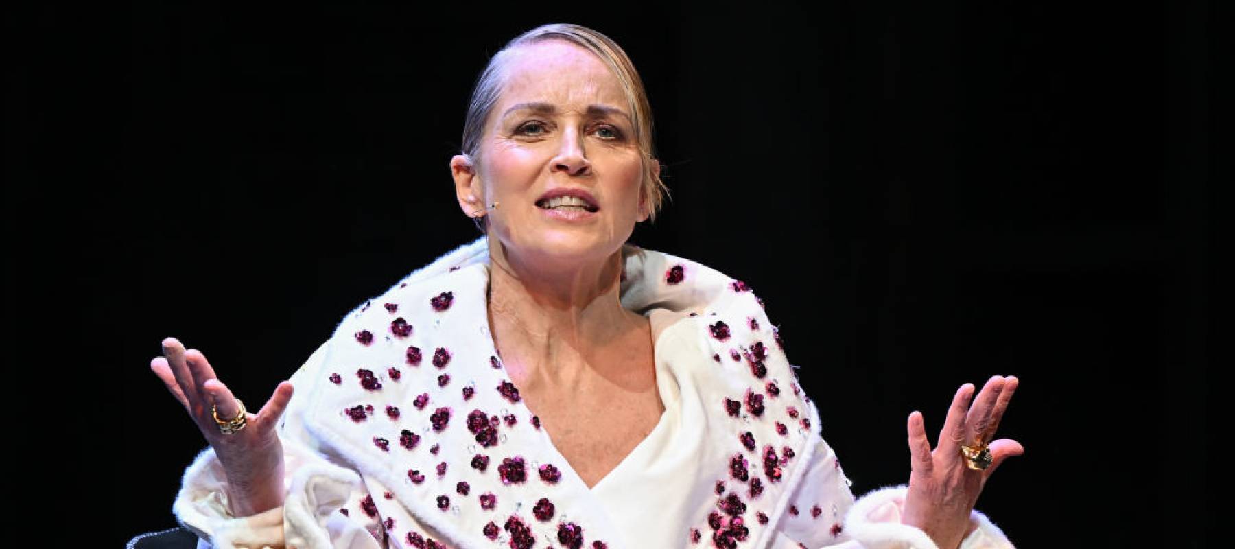 Sharon Stone on stage with an inquisitive look on her face, holding her arms up in a shrug.