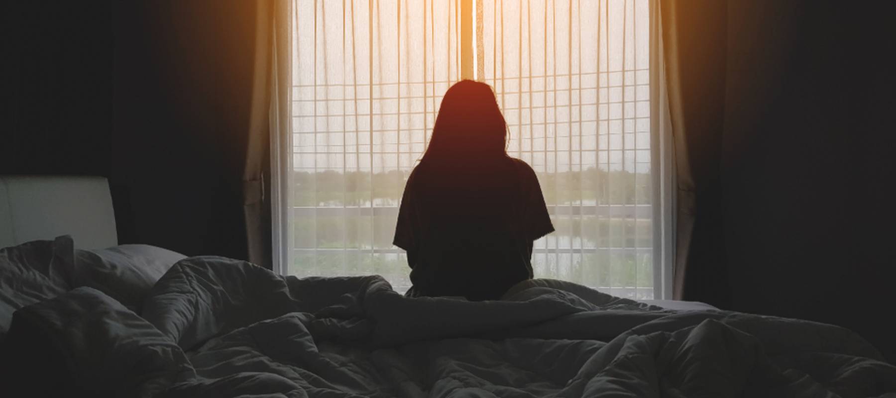 a lonely woman with her back to the camera, sitting on her bed and looking out the window