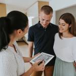 Female estate agent holding digital tablet and talking to young first time buyers in new apartment.