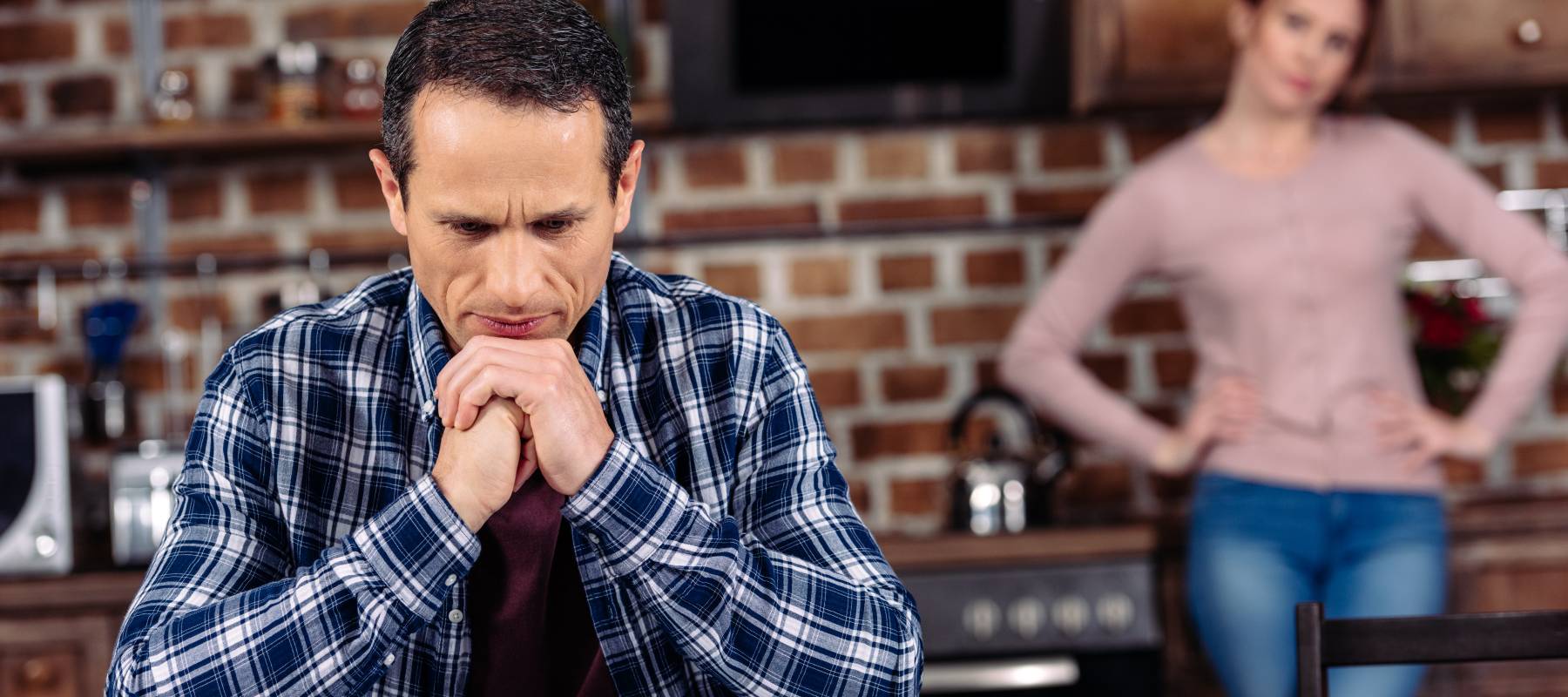 A man sits pensively at a table while his wife stands behind him.