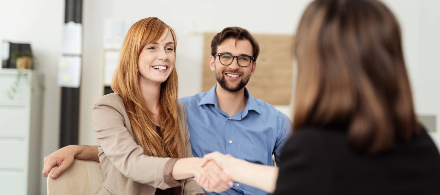 Happy young couple meeting with a broker in her office leaning over the desk to shake hands, view from behind the female agent