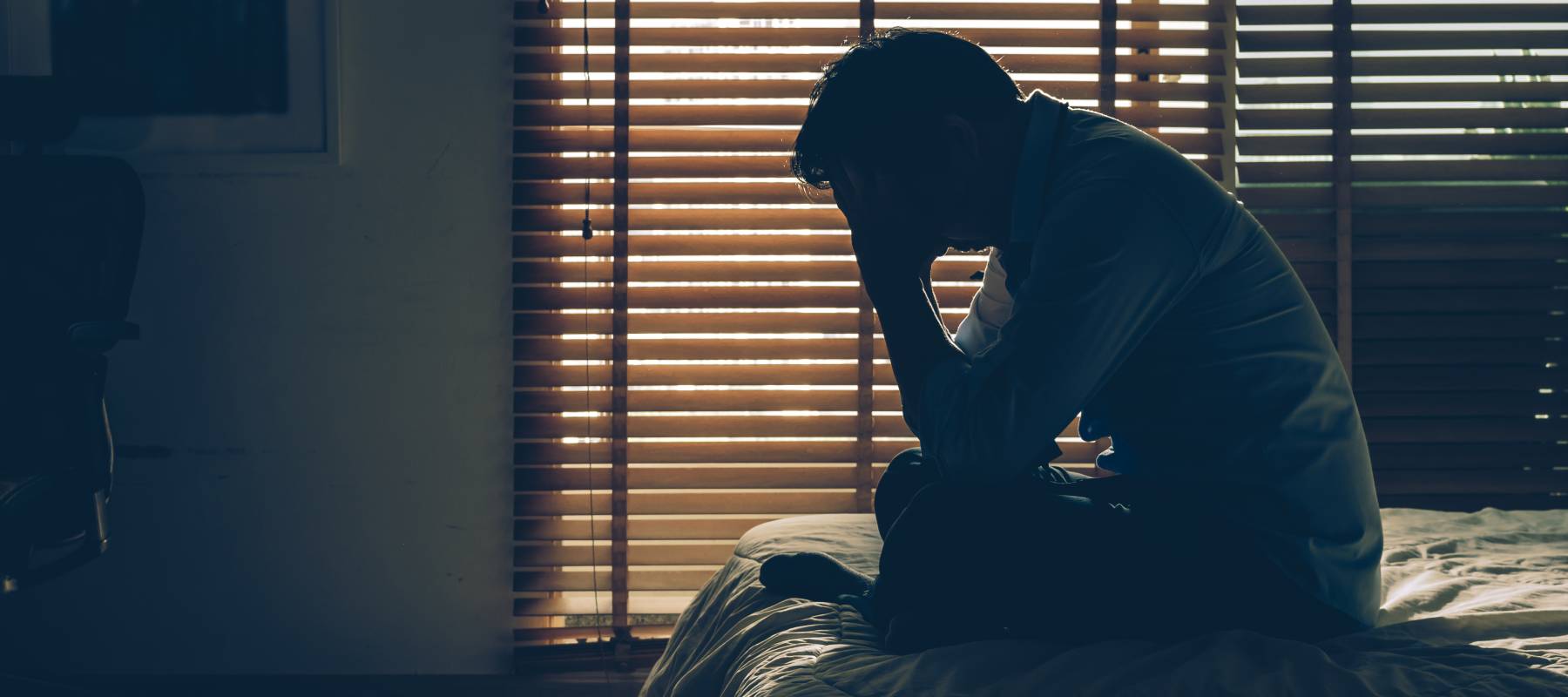silhouette of a senior man sitting on his bed in a darkened room, head in hands