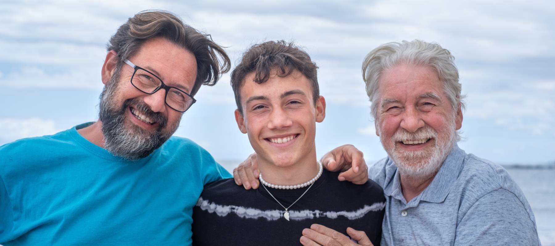 Three generations of men in same family on cloudy day, smiling at camera.