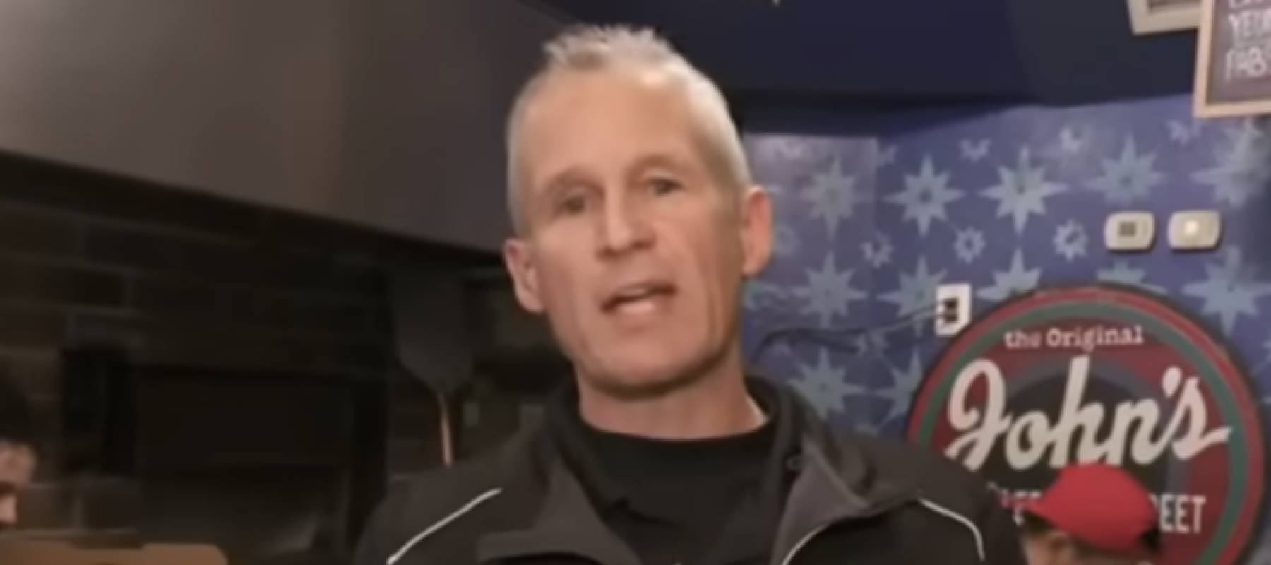 Man seen speaking to camera while pizzas are made behind him.