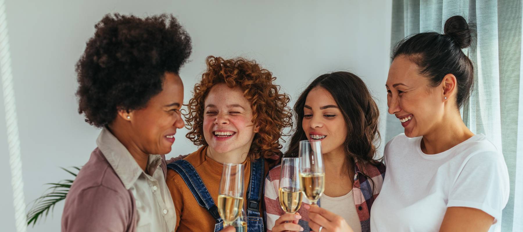 Group of women standing in a living room, arms around each other, clinking glasses and smiling.