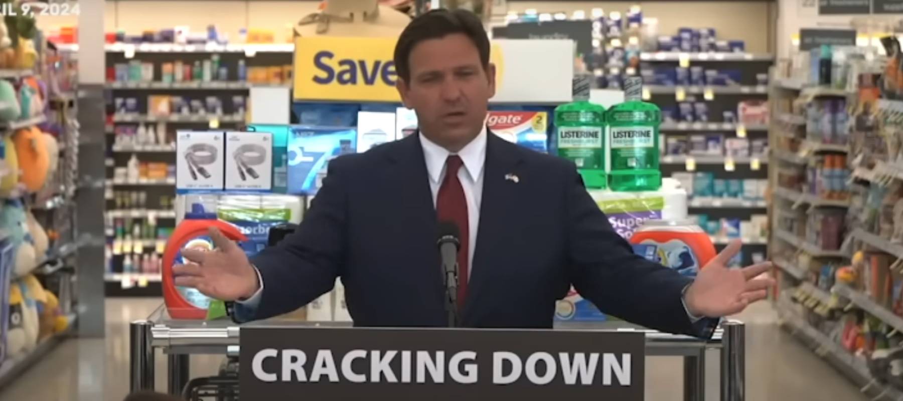 Ron DeSantis speaking with arms open behind a podium, with a retail store and supplies seen behind him.