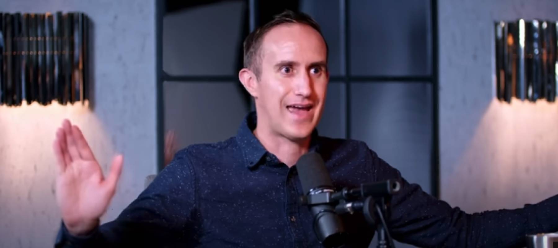 Man in blue button up shirt speaking into a microphone on a podcast set, arms stretched out and eyebrows raised in seriousness.