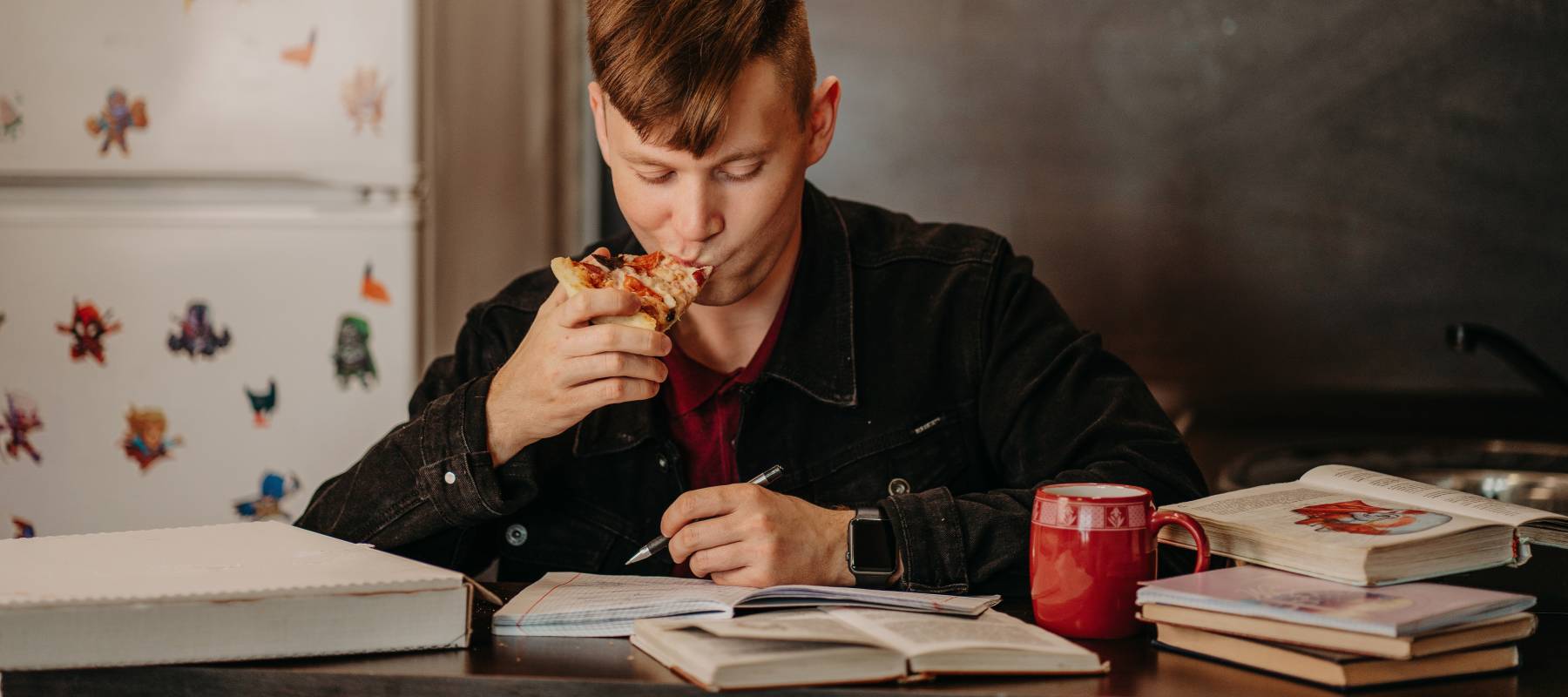 Student sitting at the table in the student dormitory eating pizza and reading textbooks.