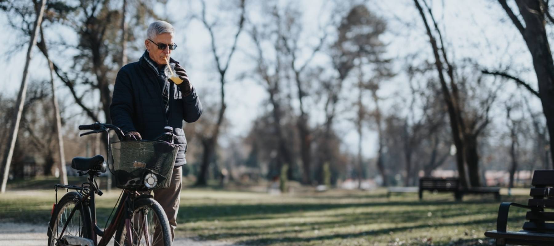 Caucasian senior man walking bike through park, drinking iced coffee