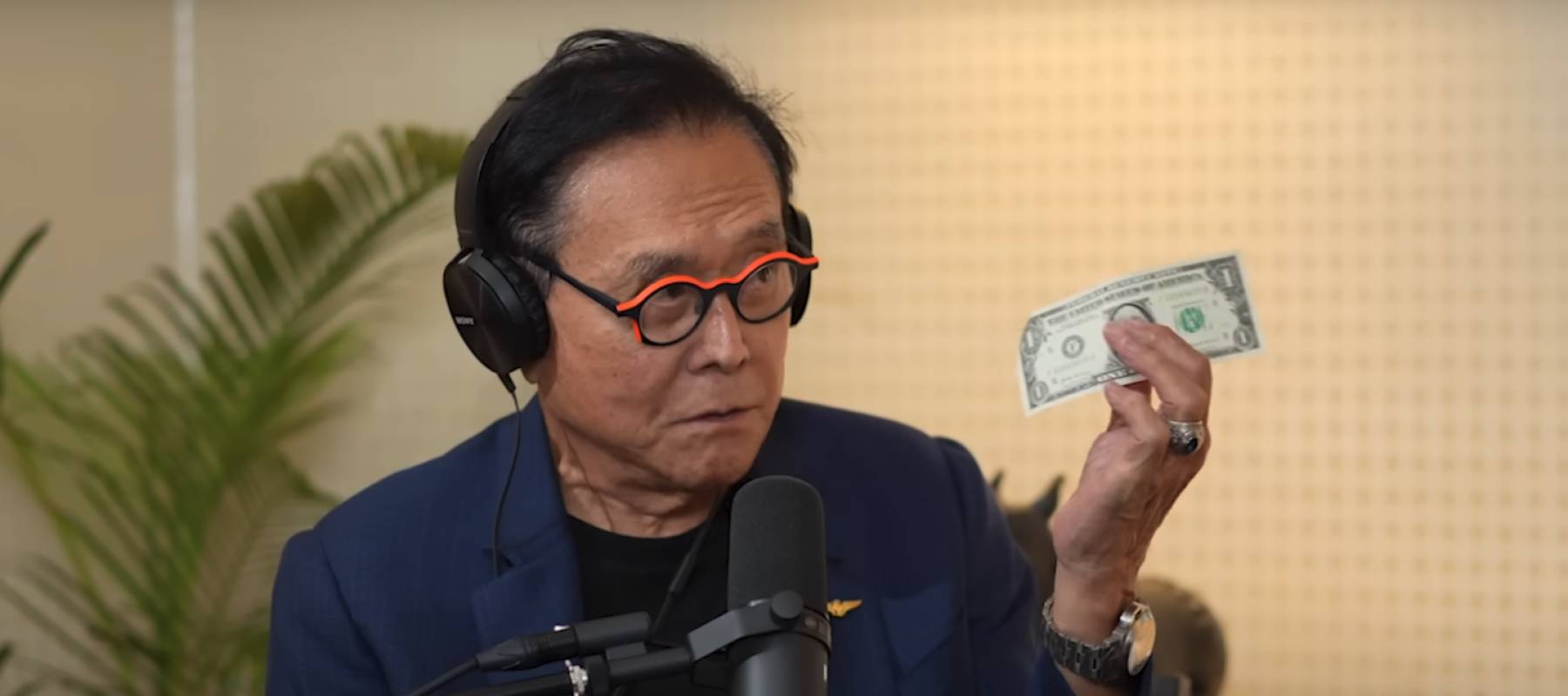 Robert Kiyosaki seen in a podcast studio holding a dollar bill up.