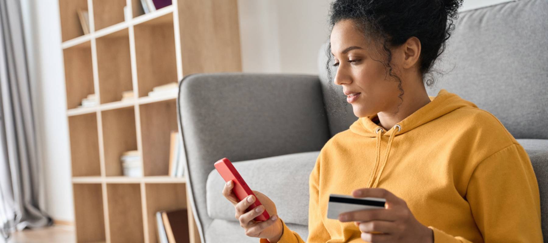 frican American female consumer holding credit card and smartphone sitting on floor at home