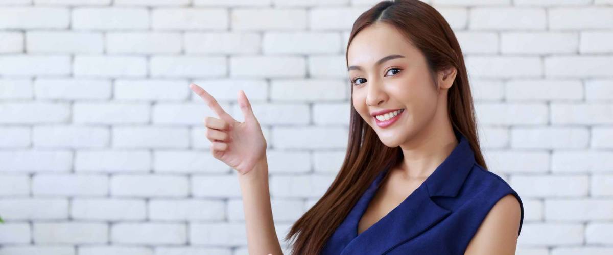 Young attractive happy Asian woman gesturing pointing at copy space, standing position at the office.