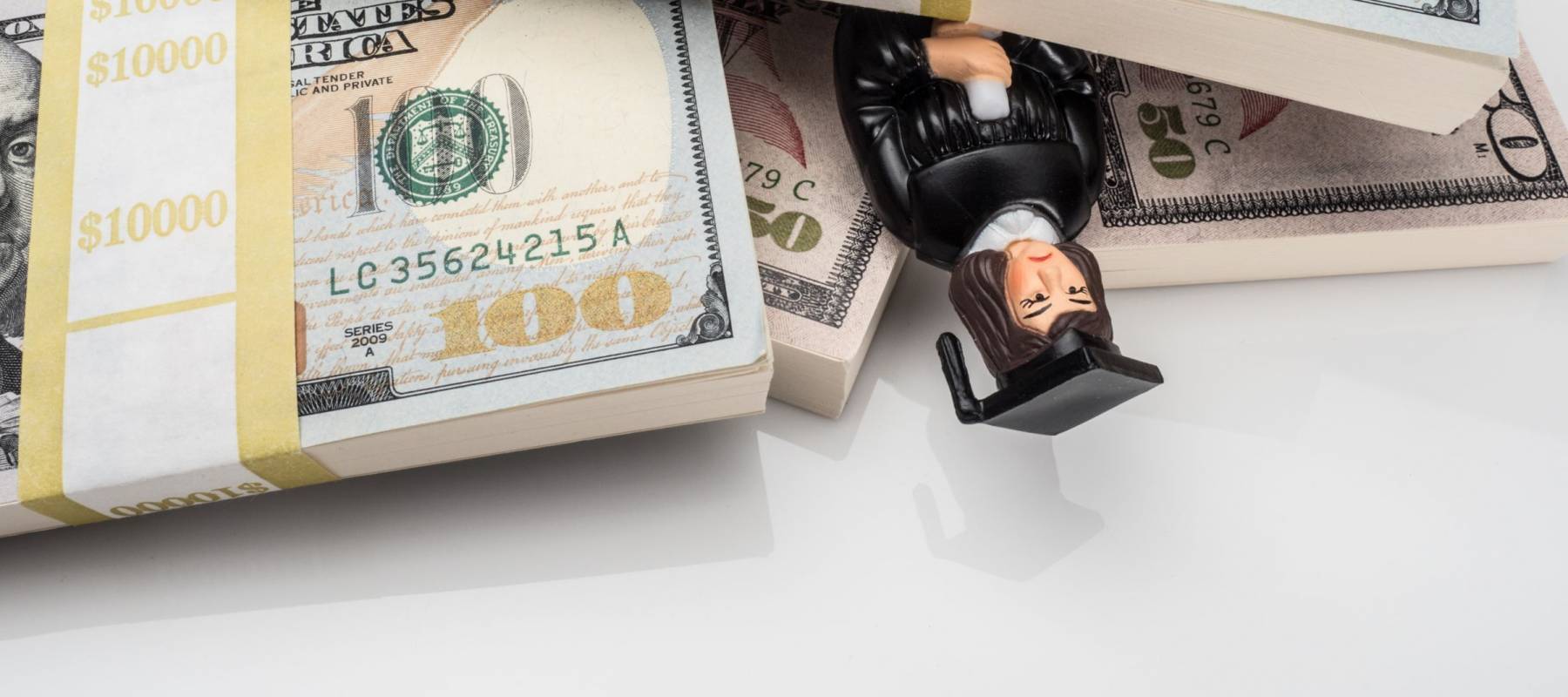 Photograph of figurine of unhappy female graduate in cap and gown buried under stacks of U.S. paper currency, suggesting the idea of the high price of education and crushing student loans.