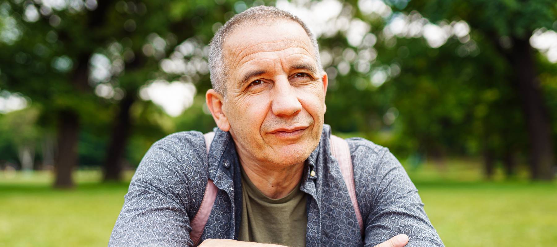 Older man sitting on picnic table in park, arms crossed and looking serious.