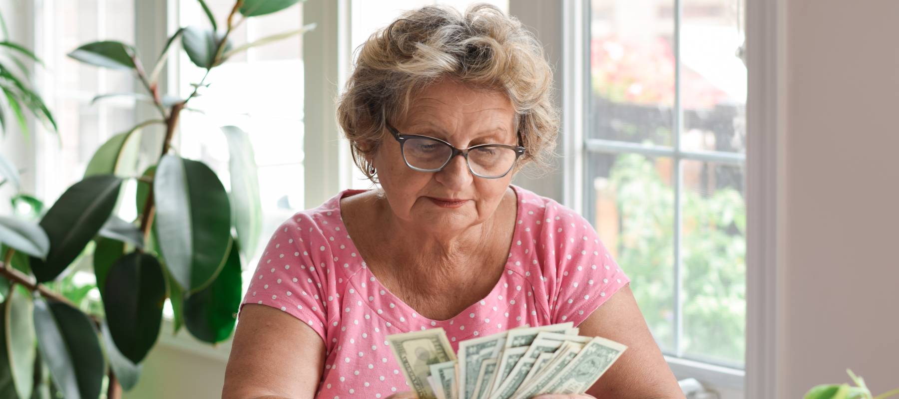 Senior woman with glasses in pink top sitting at desk at home counting American cash money dollars.