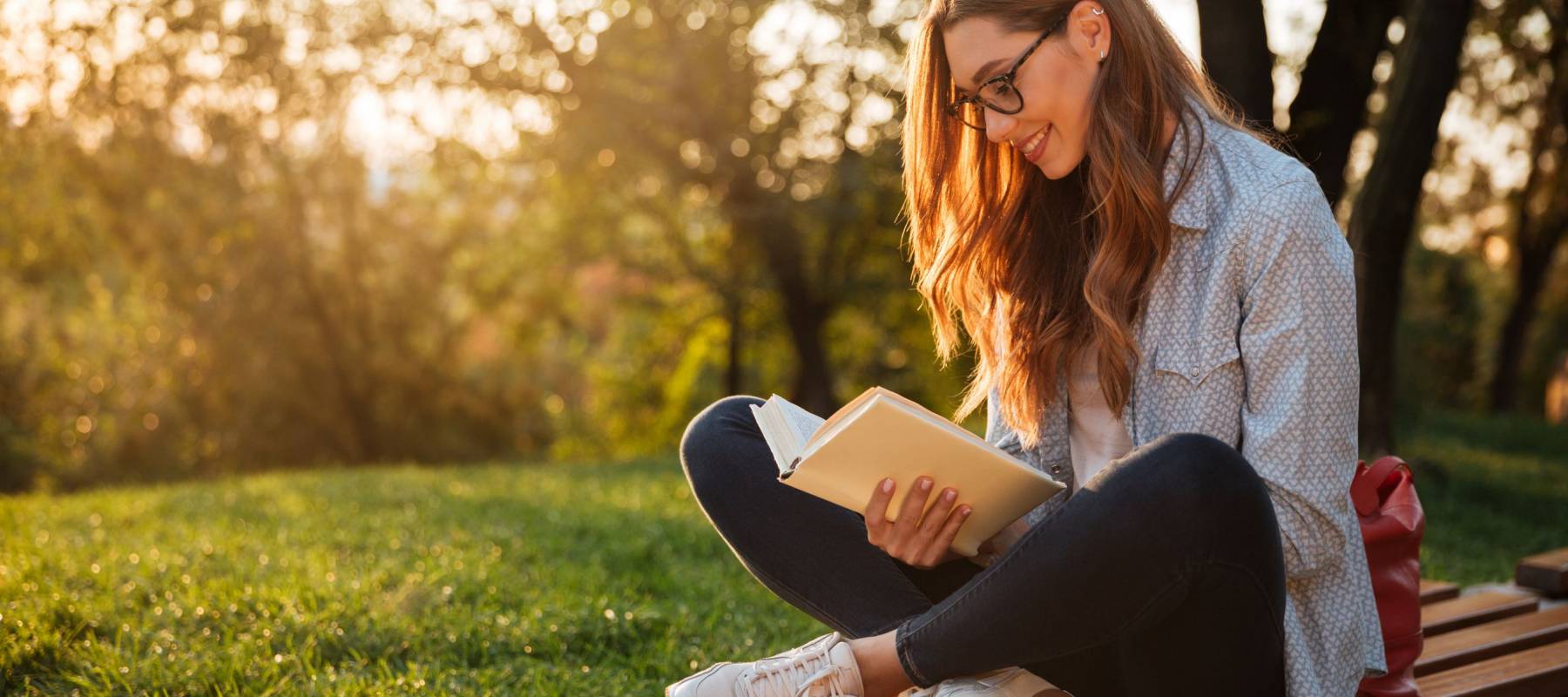 Side view of pleased brunette woman in eyeglasses sitting on bench and reading book in park