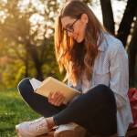 Side view of pleased brunette woman in eyeglasses sitting on bench and reading book in park