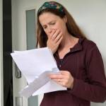 Shocked adult woman (female age 25-35) reading a letter outside a home front door.