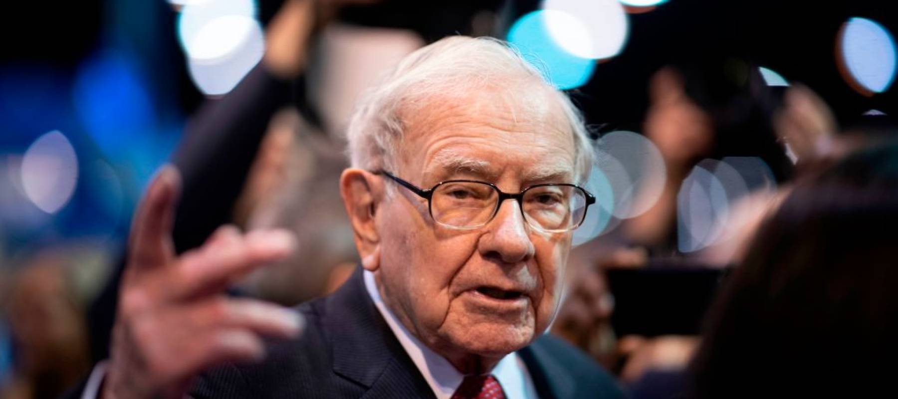 Close up of Warren Buffett, gesturing, talking in the middle of a crowd.
