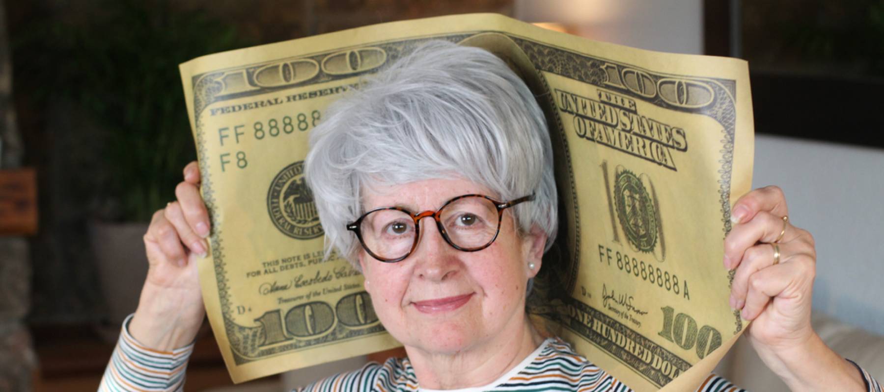 Senior woman holds giant $100 bill on her shoulders.