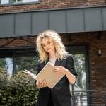 stylish blonde real estate agent looking at documents in folder near contemporary urban building