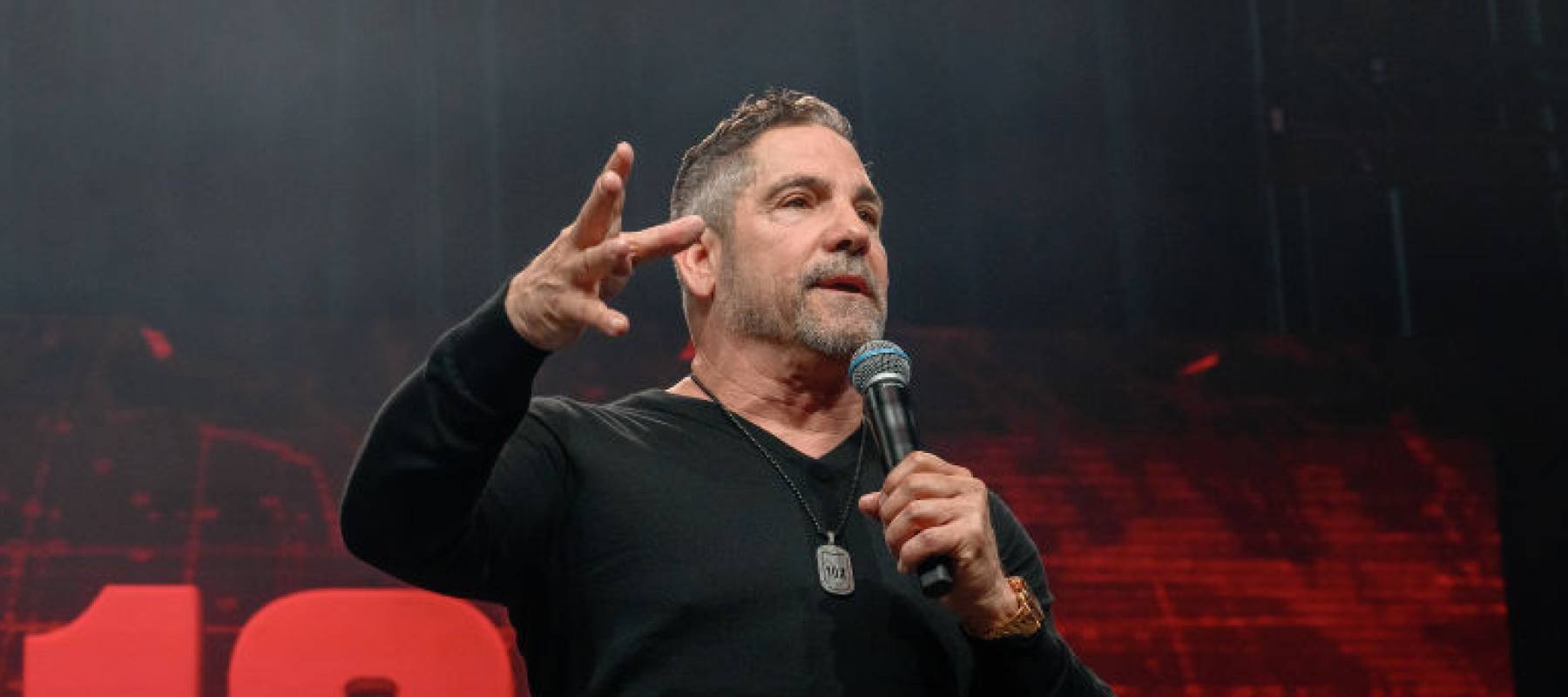 Grant Cardone seen on stage speaking into a microphone, holding out his other hand with palm facing the ground.