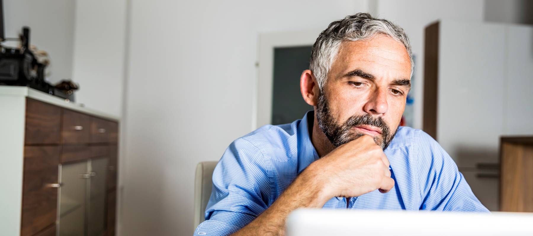 Older man working at laptop at kitchen table, looking contemplative and serious.