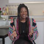 Woman sits in colorful knit cardigan in her living room, speaking with a smile and her hands held up.
