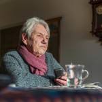 Older woman sits lost in thought with a cup of tea at her dining table.