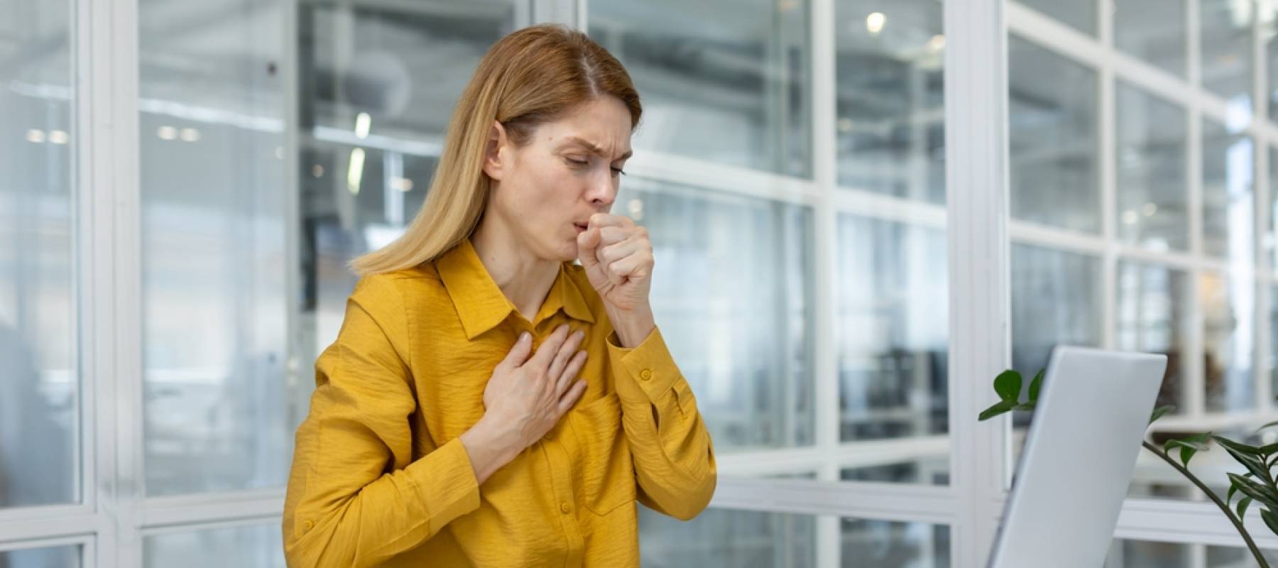 Woman experiencing a cough and chest pain while working on a laptop in a modern office environment.