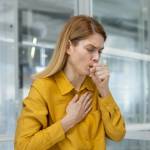 Woman experiencing a cough and chest pain while working on a laptop in a modern office environment.