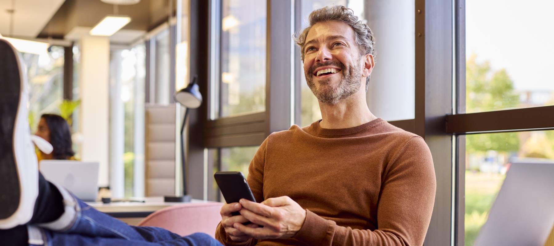 Casually Dressed Mature Businessman With Feet On Desk In Office Using Mobile Phone