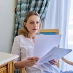 Sad emotion on face of mature woman reading paper letter, contract, document. Upset female sitting at home on chair