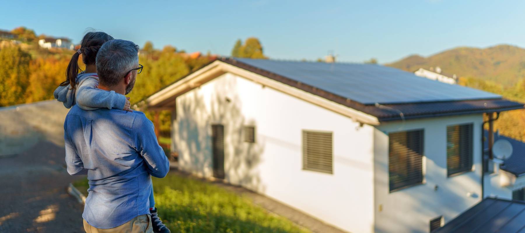 Rear view of dad holding her little girl in arms and looking at their house with solar panels.Alternative energy, saving resources and sustainable lifestyle concept.