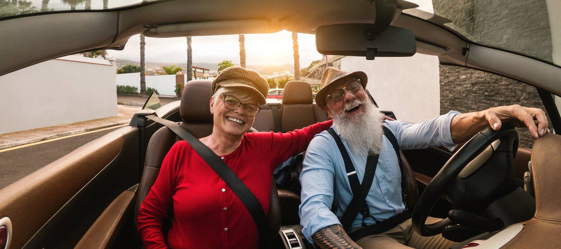 Older couple seen happily riding in a convertible with top down, palm trees and road behind them.