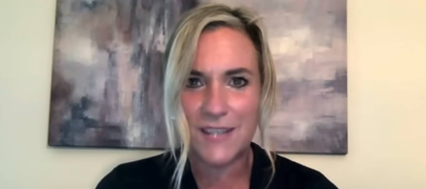 Woman seen looking serious, speaking directly to camera, sitting in front of wall.